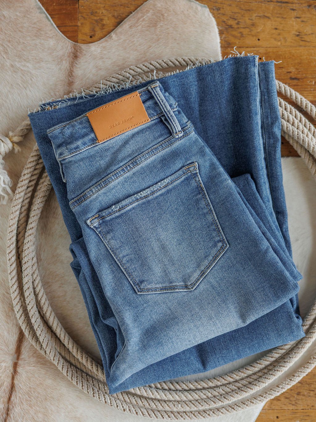 Two pairs of The Slayden High Rise Wide Leg Jeans by Savannah Sevens western life{&}style, one showing the back pocket with a brown leather patch, are neatly folded on a cowhide rug with a coiled rope around them on a wooden floor.