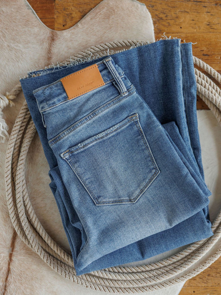 Two pairs of The Slayden High Rise Wide Leg Jeans by Savannah Sevens western life{&}style, one showing the back pocket with a brown leather patch, are neatly folded on a cowhide rug with a coiled rope around them on a wooden floor.