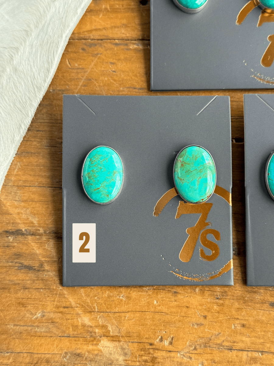 The Slidell Earrings by Savannah Sevens western life{&}style—two handmade turquoise studs in sterling silver—are shown on a gray card labeled “2” and “7s,” placed on wood with a white textured cloth partly visible in the corner.