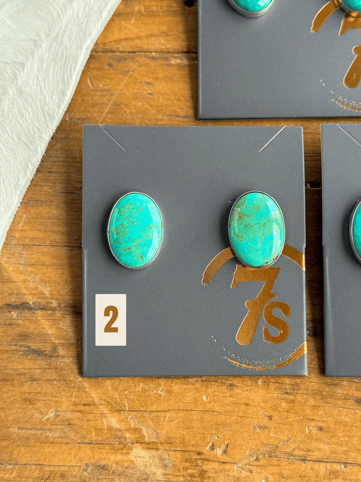 The Slidell Earrings by Savannah Sevens western life{&}style—two handmade turquoise studs in sterling silver—are shown on a gray card labeled “2” and “7s,” placed on wood with a white textured cloth partly visible in the corner.