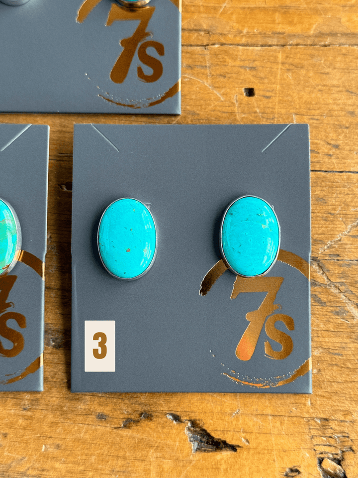 The Slidell Earrings from Savannah Sevens western life{&}style—handmade turquoise studs with sterling silver posts—are shown on a gray card featuring the gold 7s logo, labeled 3, and placed on a wooden surface.