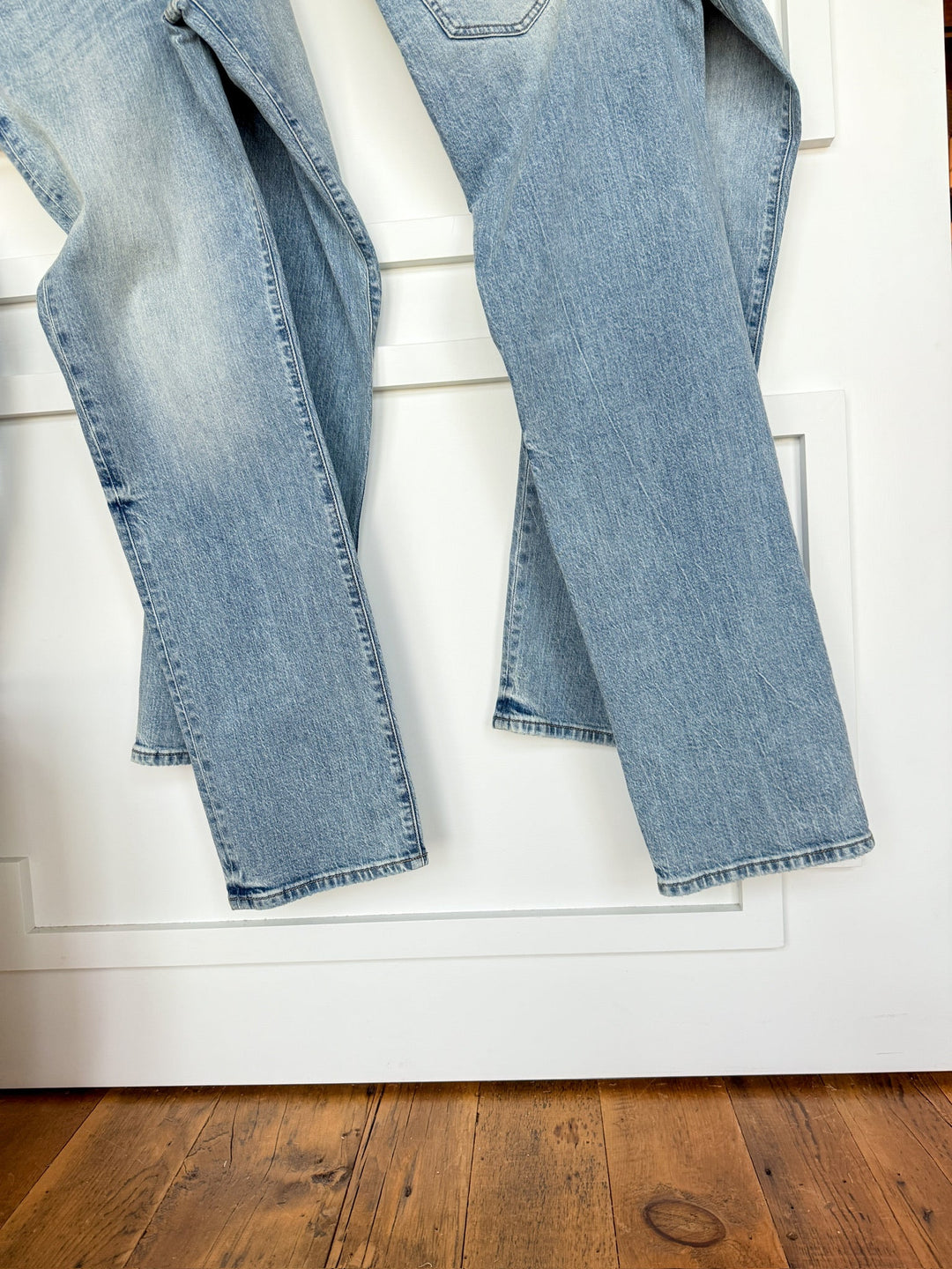 The Sloane High Rise Straight Leg Jean by Savannah Sevens western life{&}style in light wash denim hangs over a white door with decorative molding, with the wooden floor visible below.