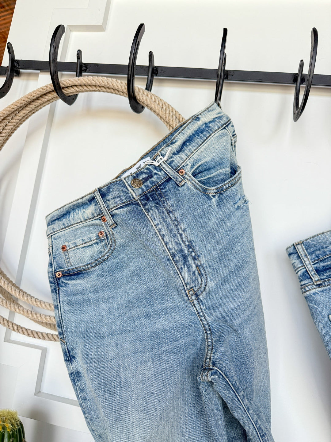The Sloane High Rise Straight Leg Jean by Savannah Sevens western life{&}style, in a light wash, hangs by the belt loop on a black hook rack with rope detail, set against a white wall.