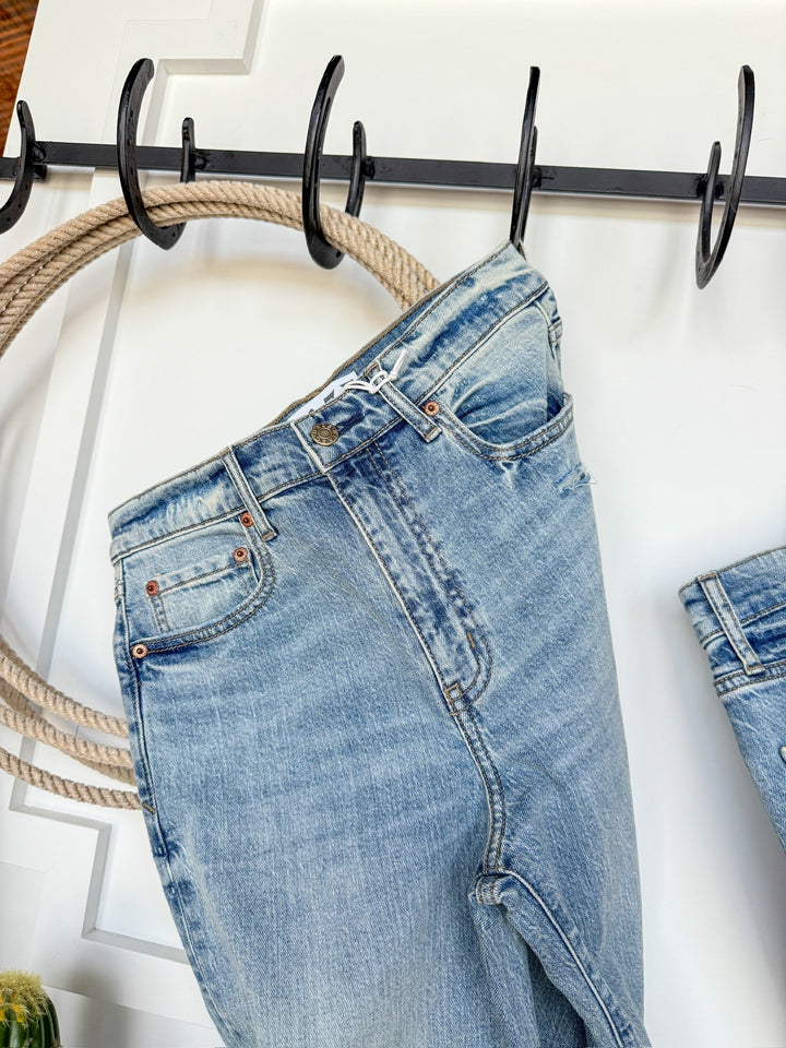 The Sloane High Rise Straight Leg Jean by Savannah Sevens western life{&}style, in a light wash, hangs by the belt loop on a black hook rack with rope detail, set against a white wall.