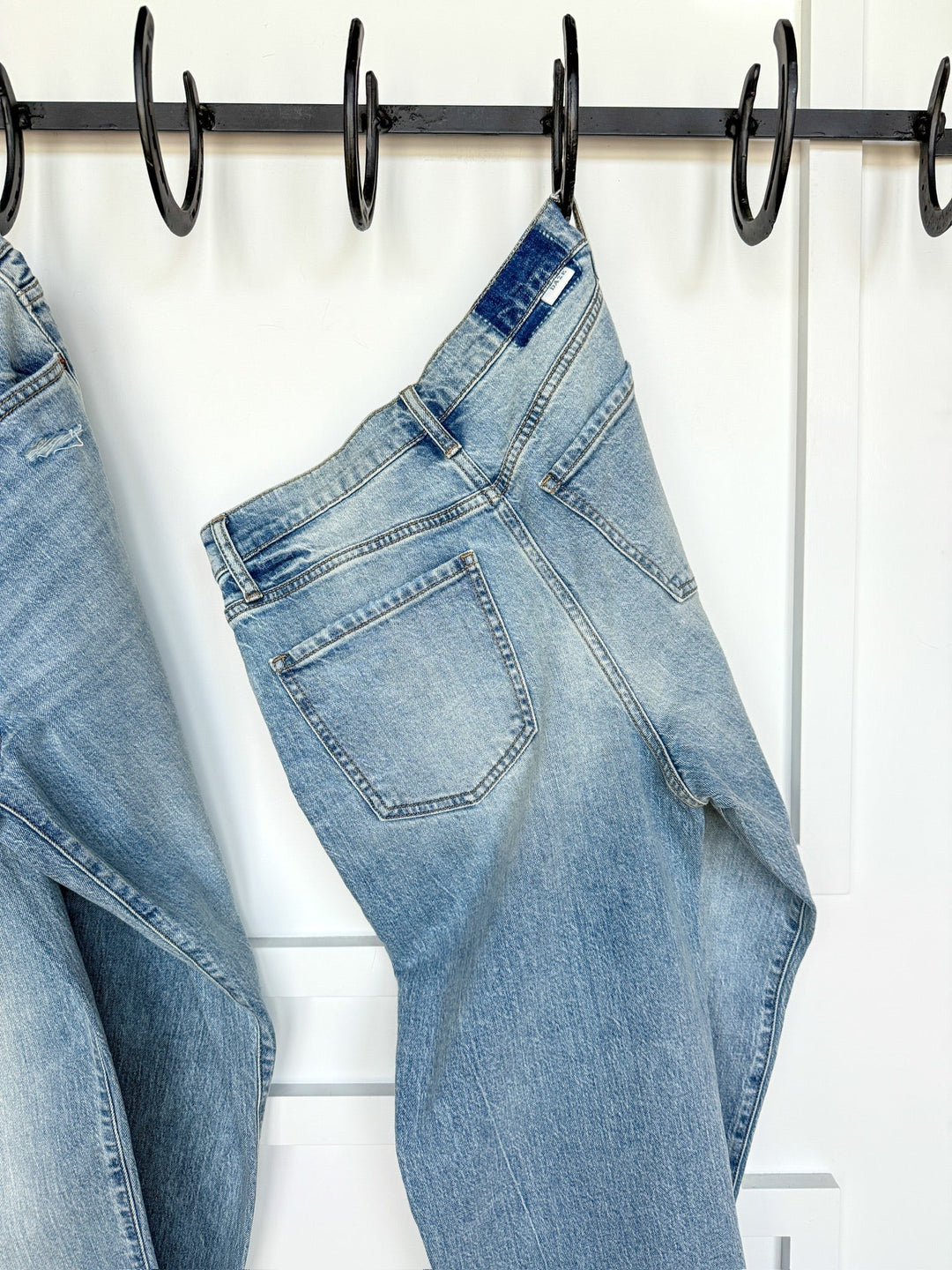 The Sloane High Rise Straight Leg Jean by Savannah Sevens western life{&}style in light wash hangs on a black hook against a white wall, with another pair partially visible on the left.