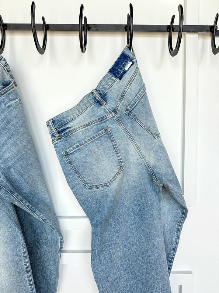 The Sloane High Rise Straight Leg Jean by Savannah Sevens western life{&}style in light wash hangs on a black hook against a white wall, with another pair partially visible on the left.