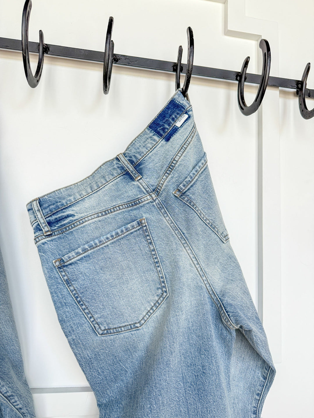 The Sloane High Rise Straight Leg Jean by Savannah Sevens western life{&}style in light wash denim hangs by a belt loop on a black metal hook against a white paneled wall.