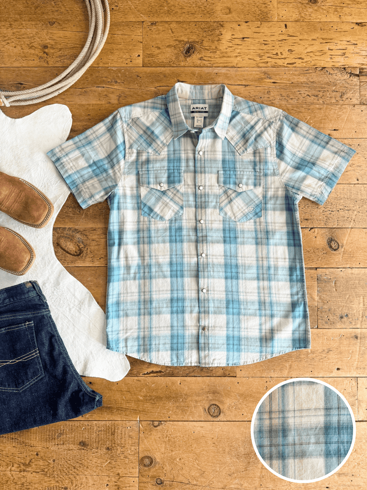 The Smithville Ariat Men's Blue Plaid Pearl Snap by Savannah Sevens western life{&}style is laid flat next to blue jeans, brown boots, a white rug, and rope; a zoomed-in circle highlights the shirt’s fabric pattern.