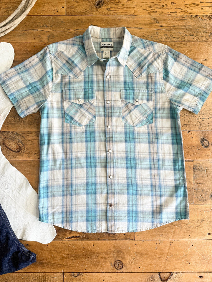 The Smithville Ariat Men's Blue Plaid Pearl Snap by Savannah Sevens western life{&}style features short sleeves, light blue and white plaid, two front chest pockets, and a classic western retro fit, shown flat on a wooden surface.