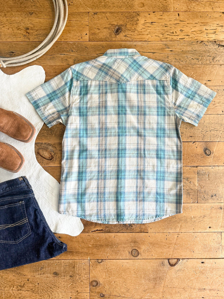 The Smithville Ariat Men's Blue Plaid Pearl Snap from Savannah Sevens western life{&}style is shown laid flat on a wooden floor with a white cowhide rug, brown boots, and dark blue jeans—an ideal men's western shirt with retro fit style.