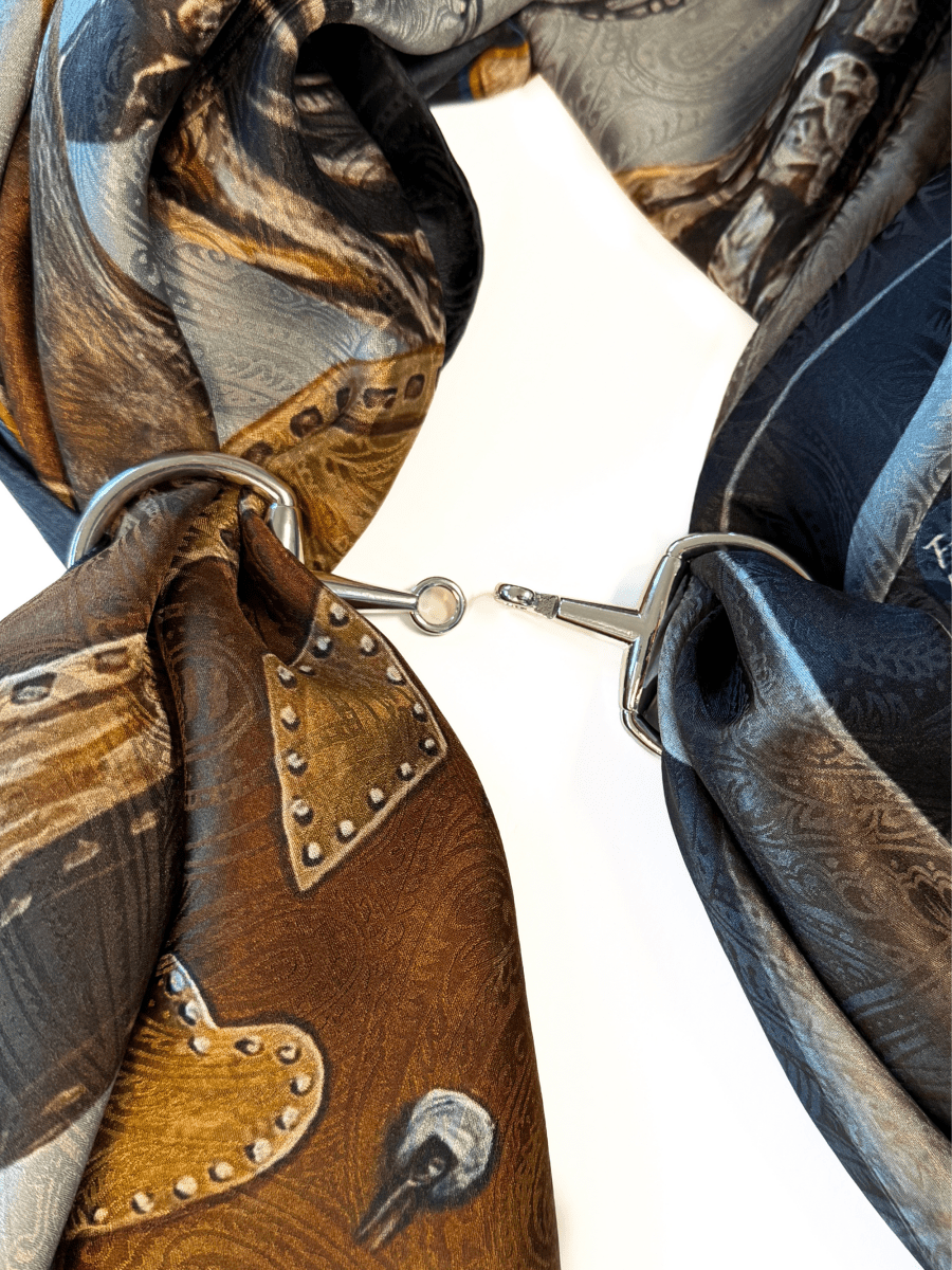 A close-up of two patterned silk scarves looped through the Savannah Sevens western life{&}style Snaffle Bit Scarf Buckle, joined in the center by a small metal hook, set on a white background.