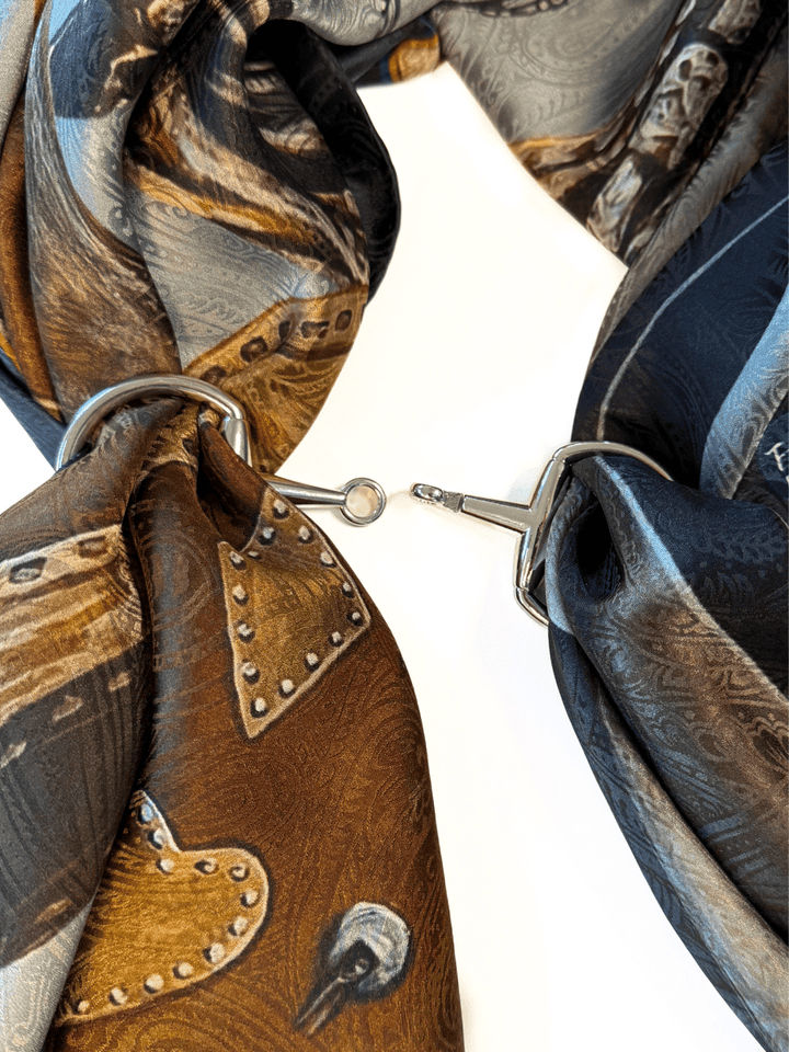 A close-up of two patterned silk scarves looped through the Savannah Sevens western life{&}style Snaffle Bit Scarf Buckle, joined in the center by a small metal hook, set on a white background.