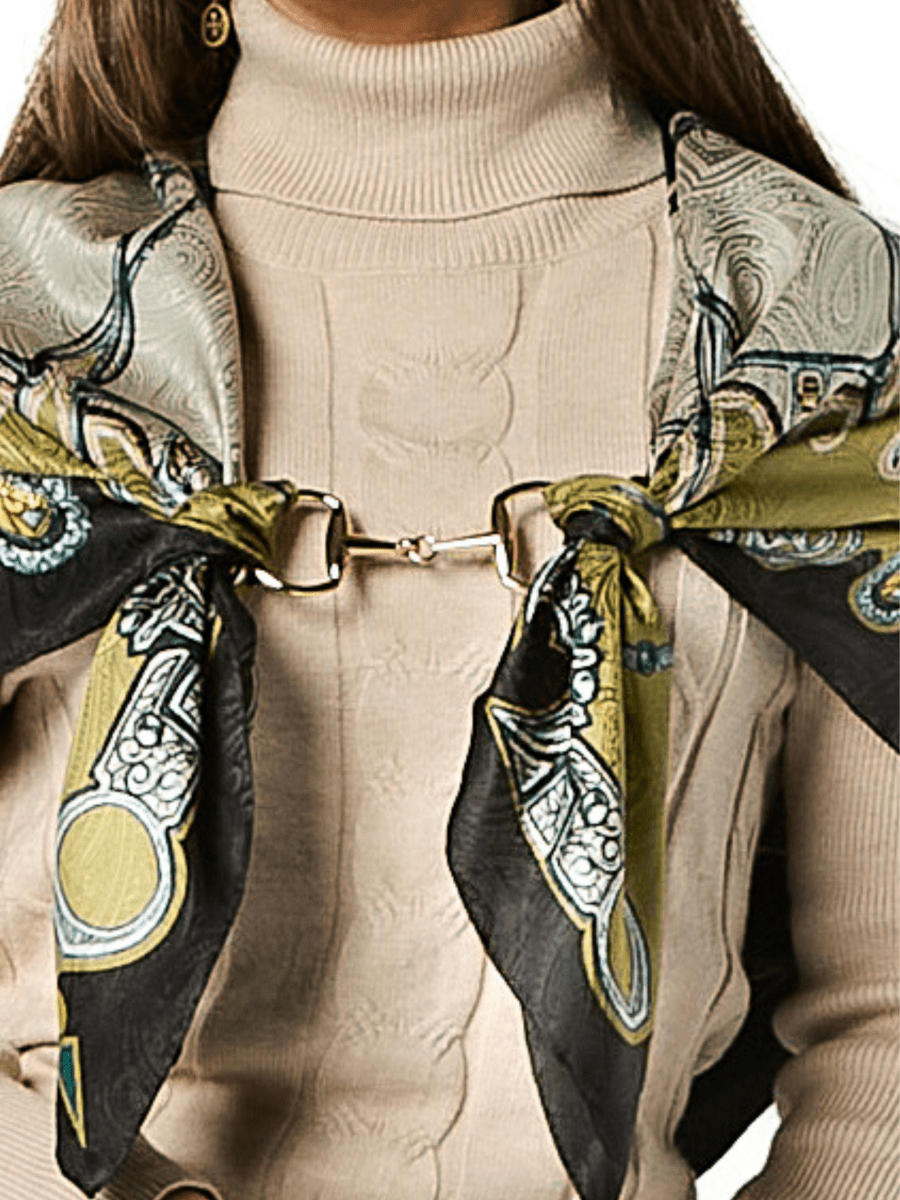 A person styles a patterned silk scarf, featuring green, black, and cream designs, at the chest with the Savannah Sevens western life{&}style Snaffle Bit Scarf Buckle over a beige cable-knit turtleneck sweater.