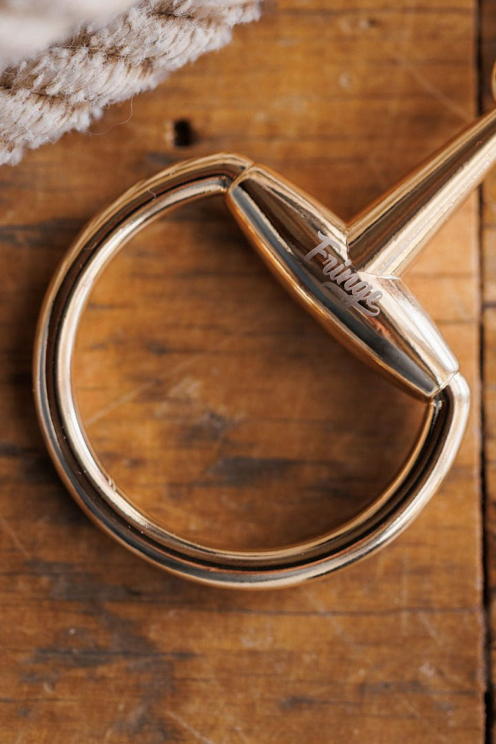 A close-up of the Savannah Sevens western life{&}style Snaffle Bit Scarf Buckle, metal with "Trust" engraved, attached to a handle and resting on wood with textured fabric in the corner.