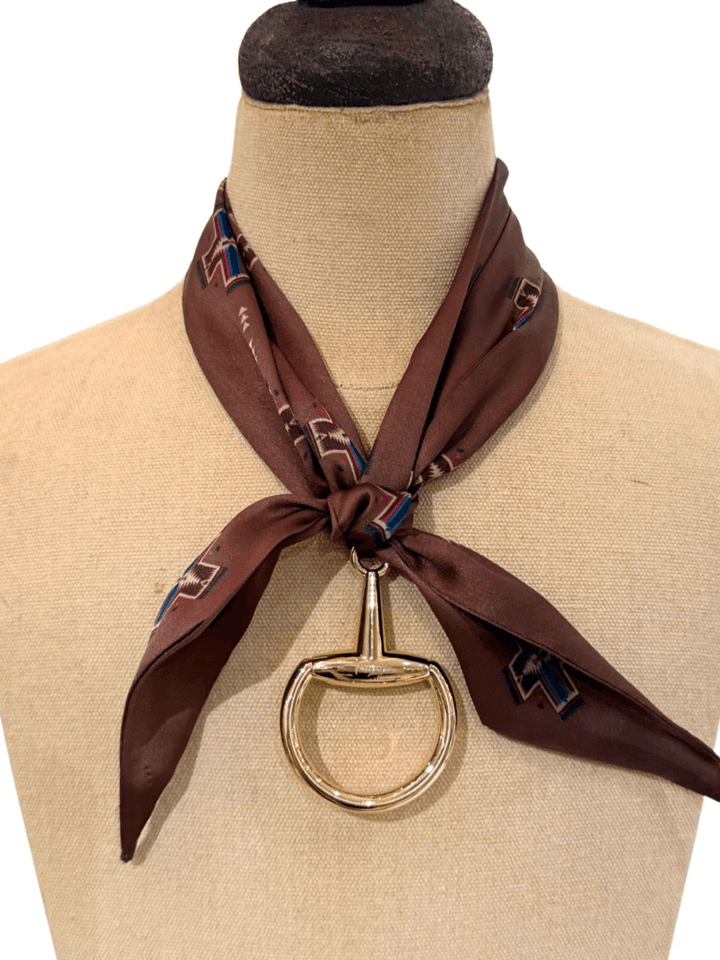 A brown silk scarf with a geometric pattern is styled on a dress form’s neck and fastened at the front using the Savannah Sevens western life{&}style Snaffle Bit Scarf Buckle for a bold statement look.