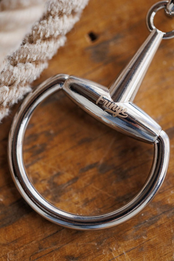 A close-up of a shiny metallic keyring with "Savannah Sevens western life{&}style" engraved, resting on a wooden surface next to textured beige fabric and a Snaffle Bit Scarf Buckle.