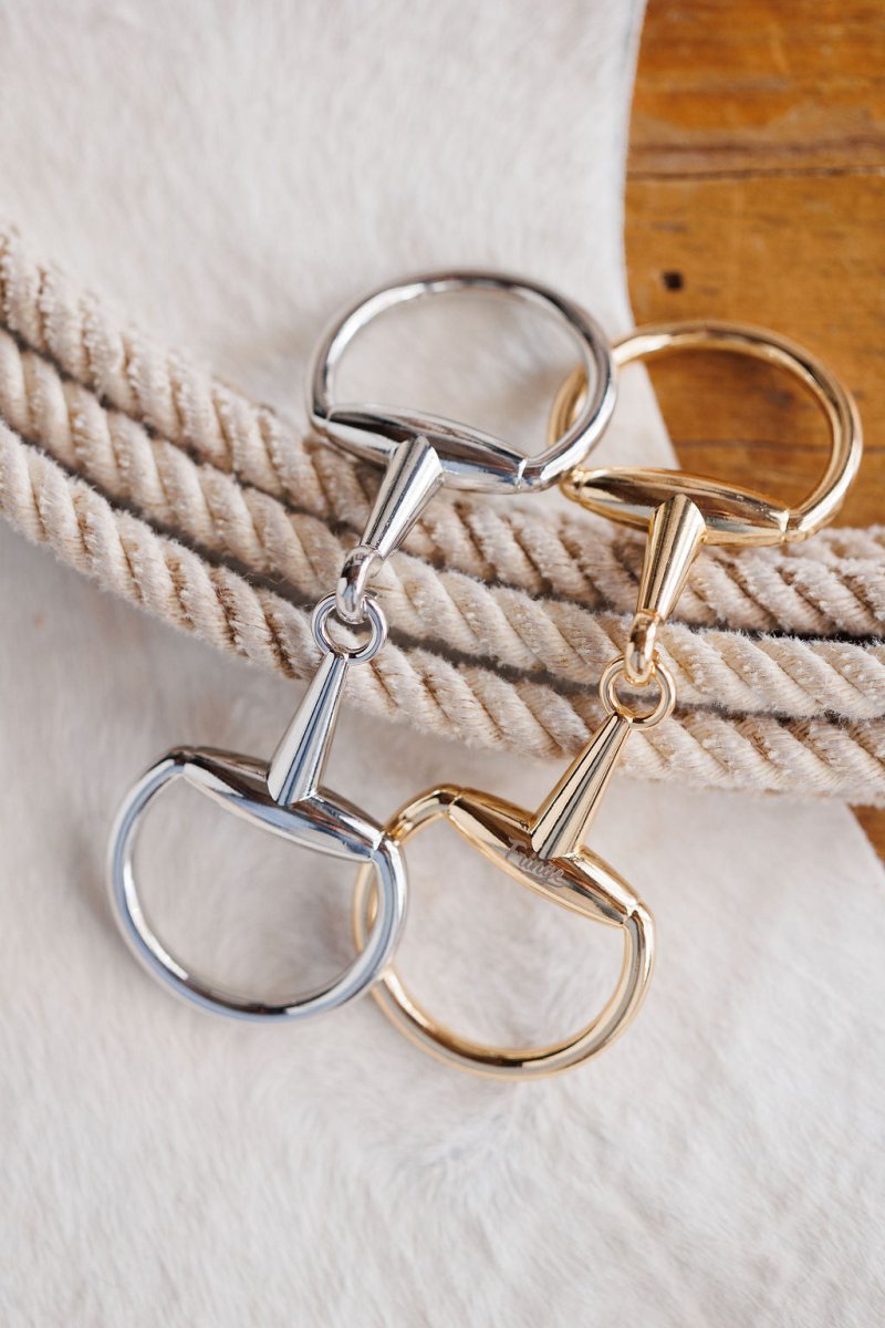 Two Snaffle Bit Scarf Buckles by Savannah Sevens western life{&}style, in silver and gold tones, are displayed on coiled beige rope atop a white surface and a wooden table.