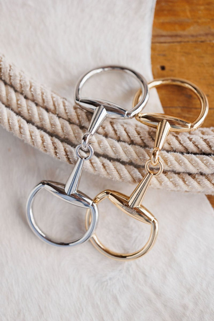 Two Snaffle Bit Scarf Buckles by Savannah Sevens western life{&}style, in silver and gold tones, are displayed on coiled beige rope atop a white surface and a wooden table.