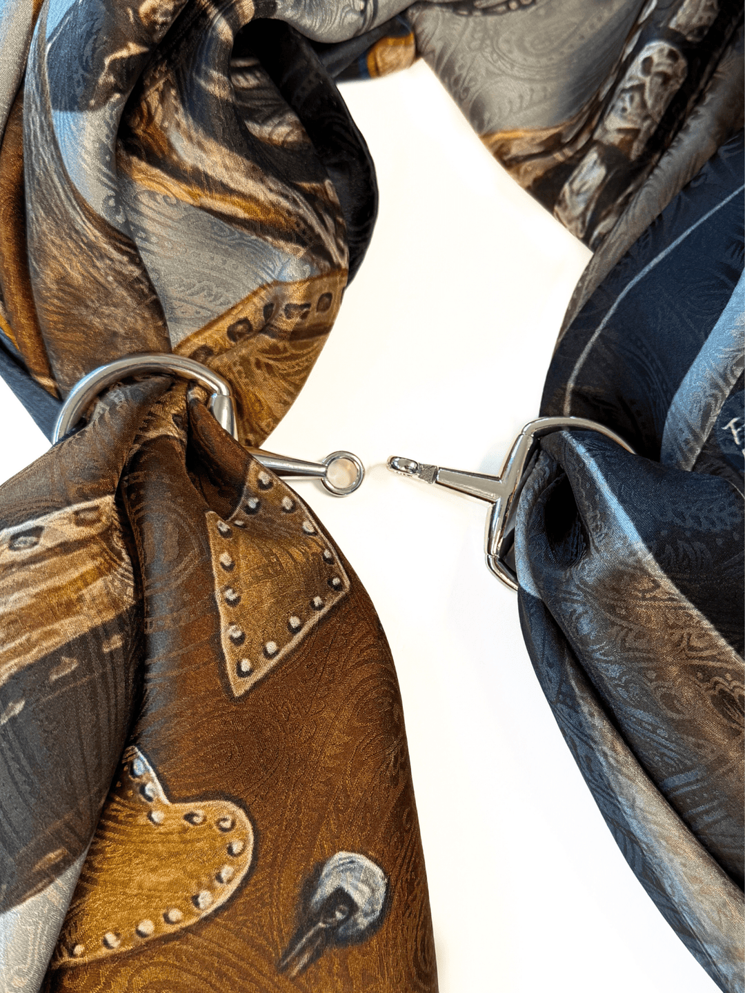 A close-up of two patterned silk scarves looped through the Savannah Sevens western life{&}style Snaffle Bit Scarf Buckle, joined in the center by a small metal hook, set on a white background.