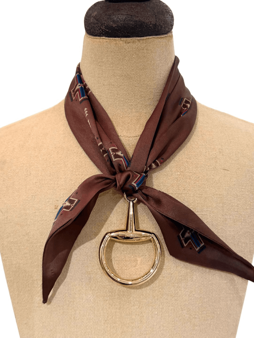 A brown silk scarf with a geometric pattern is styled on a dress form’s neck and fastened at the front using the Savannah Sevens western life{&}style Snaffle Bit Scarf Buckle for a bold statement look.
