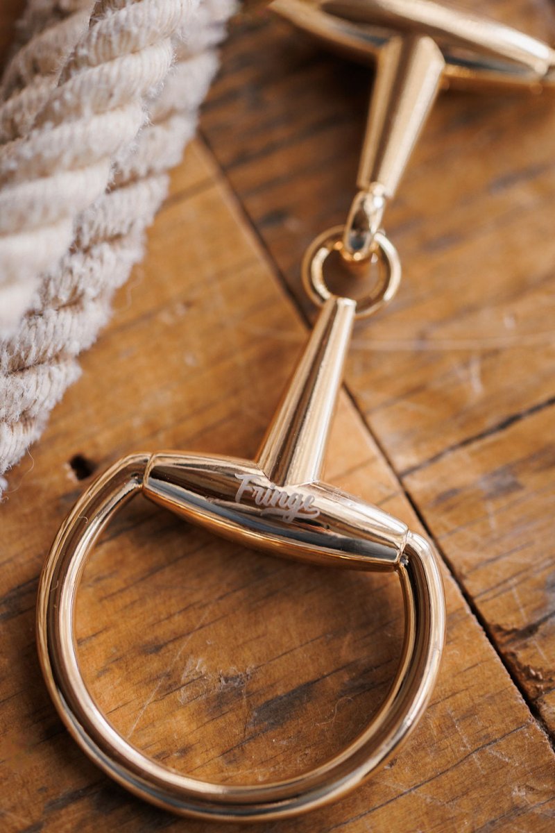 A Snaffle Bit Scarf Buckle from Savannah Sevens western life{&}style rests on a wooden surface beside a coiled rope and a statement tie.