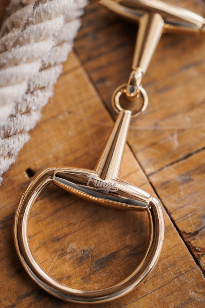 A Snaffle Bit Scarf Buckle from Savannah Sevens western life{&}style rests on a wooden surface beside a coiled rope and a statement tie.
