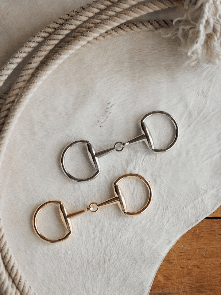 The Snaffle Bit Scarf Buckle by Savannah Sevens western life{&}style features both silver and gold metal bits on a white hide, next to coiled ropes and wood—capturing classic Western elegance.