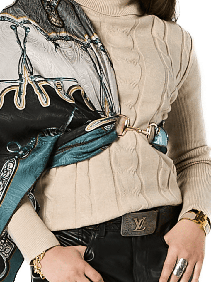 A person wears a beige cable-knit turtleneck, black leather pants, a black Louis Vuitton belt, and jewelry. A patterned silk scarf is draped over one shoulder and secured with the Savannah Sevens Snaffle Bit Scarf Buckle in gold.