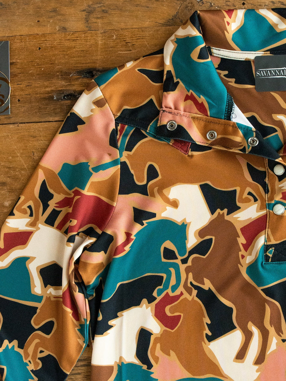 The Socorro Western Graphic Top by Savannah Sevens western life{&}style is a long sleeve pearl snap shirt showcasing a colorful horse pattern in brown, teal, cream, and black hues, partially folded on a wooden surface with metal snaps at the collar.