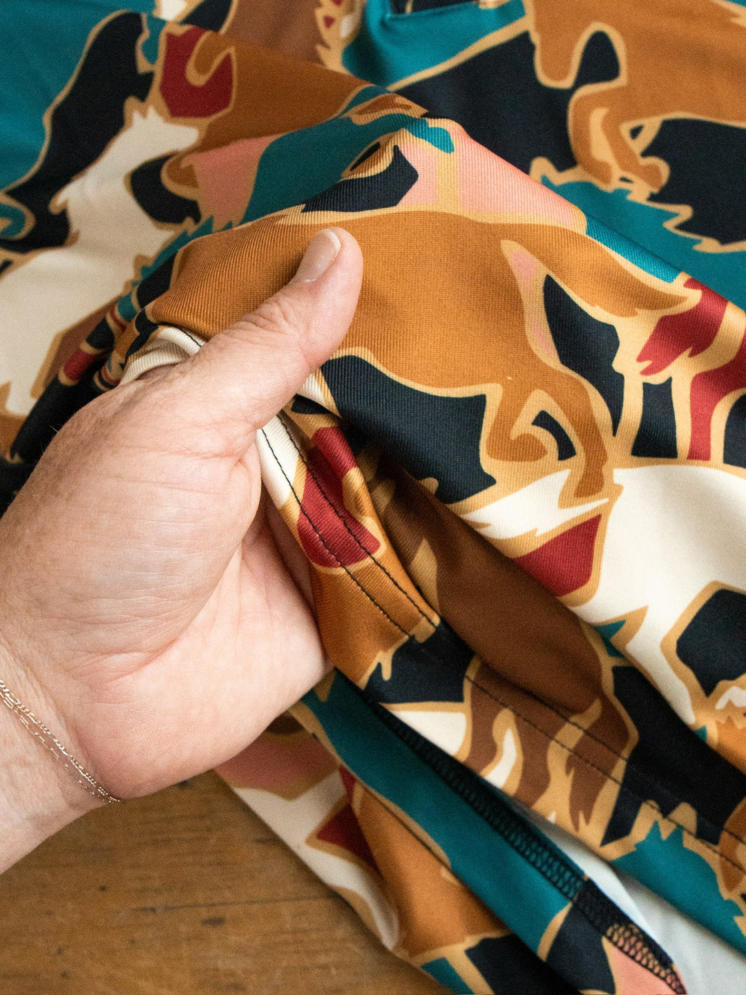 A hand displays the Socorro Western Graphic Top by Savannah Sevens western life{&}style, featuring colorful abstract patterns in shades of brown, teal, black, red, and cream on a wooden surface.