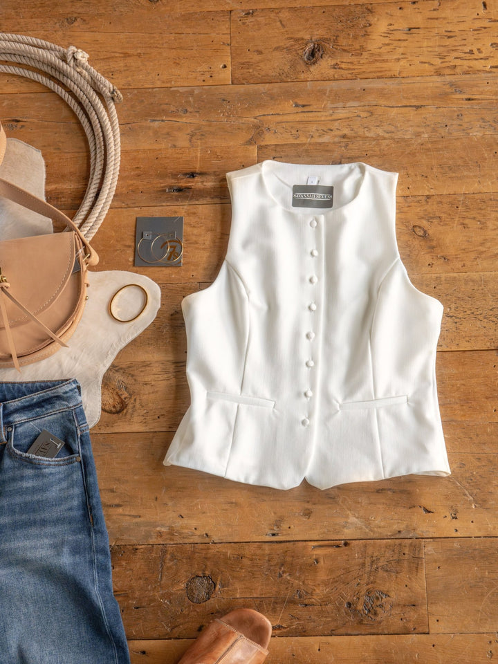 The Solano by Savannah Sevens western life{&}style, a white button-up vest, is displayed on wood with blue jeans, a tan crossbody bag, gold bangle, hoop earrings, rope, and tan sandals—this women's contemporary vest brings a modern edge to any look.