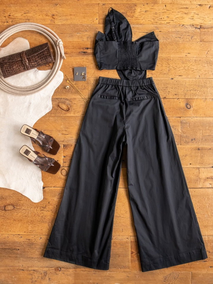 A flat lay of The Solvay {Set} by Savannah Sevens western life{&}style—a black one shoulder crop top with high waisted pants—styled with clear-heeled sandals, a brown textured clutch, gold hoop earrings, and a white rug on a wooden floor.