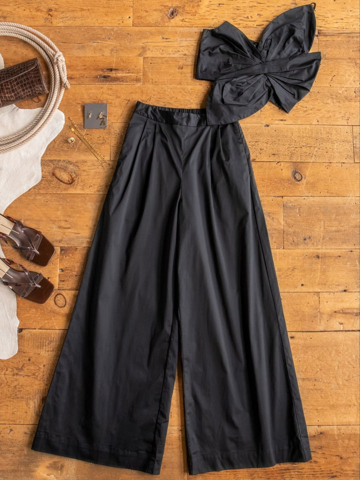 A flat lay of The Solvay Set by Savannah Sevens western life{&}style: a strapless black crop top with a large bow and high waisted pants, styled on a wooden floor with brown heeled sandals, a white bag, and jewelry.
