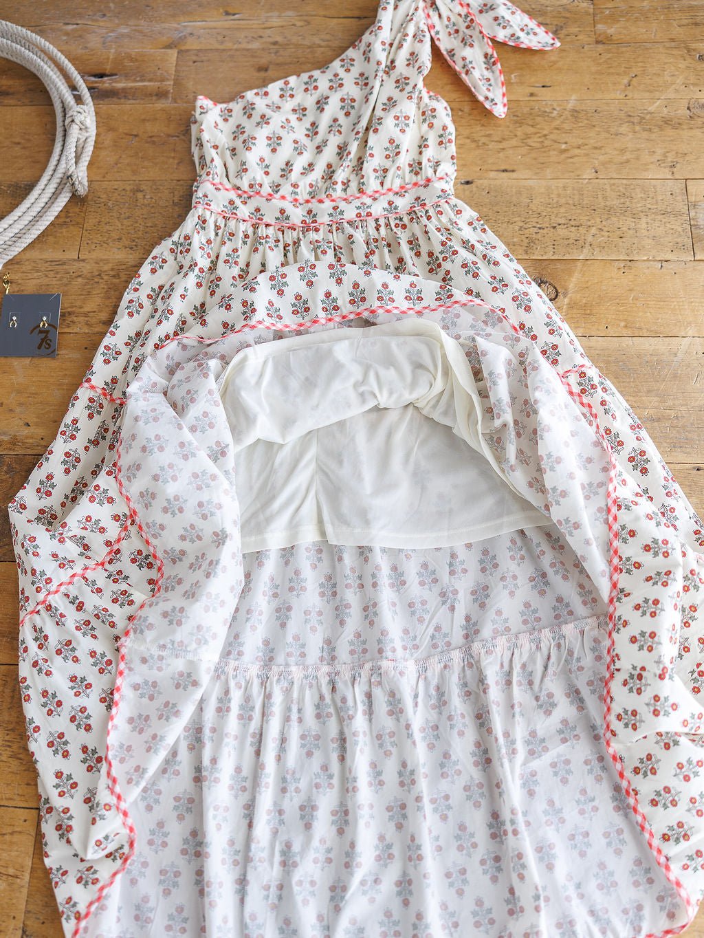 The Somerset Floral One Shoulder Dress by Savannah Sevens western life{&}style, featuring a tied strap and gingham trim, is displayed on a wooden floor with its skirt flipped up to show the white lining. A rope and black card are placed beside it.