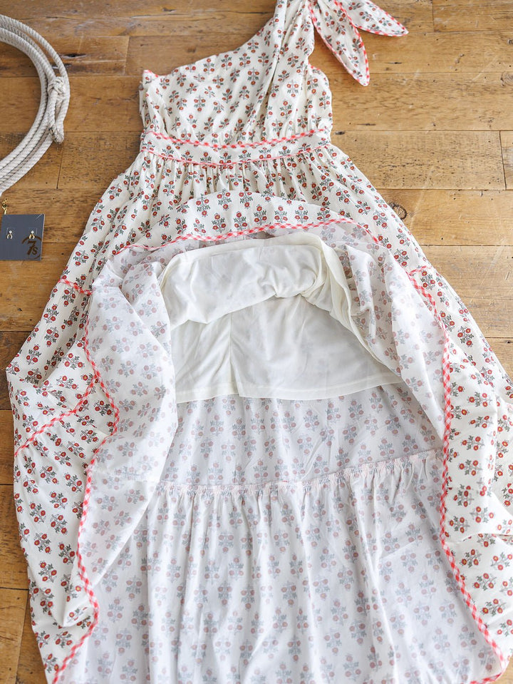 The Somerset Floral One Shoulder Dress by Savannah Sevens western life{&}style, featuring a tied strap and gingham trim, is displayed on a wooden floor with its skirt flipped up to show the white lining. A rope and black card are placed beside it.