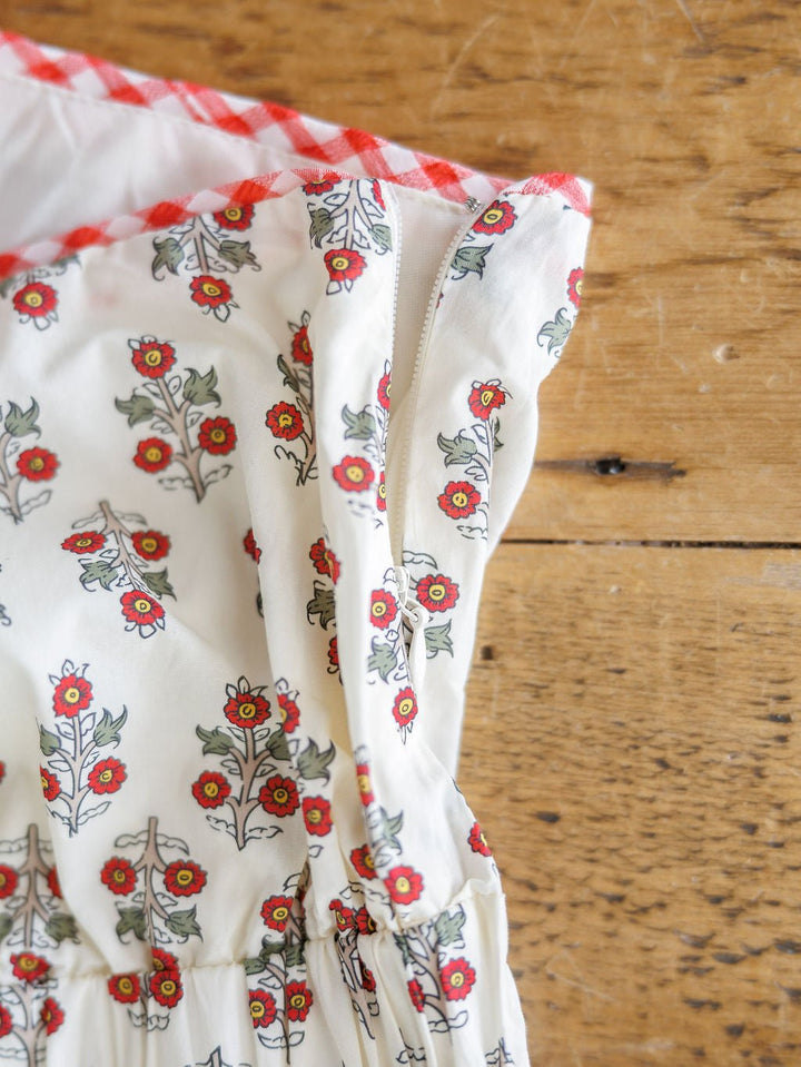 A close-up of the Somerset Floral One Shoulder Dress by Savannah Sevens western life{&}style, a cotton midi with a red floral pattern, elastic gathering, and gingham trim along the top edge, laid on wood.