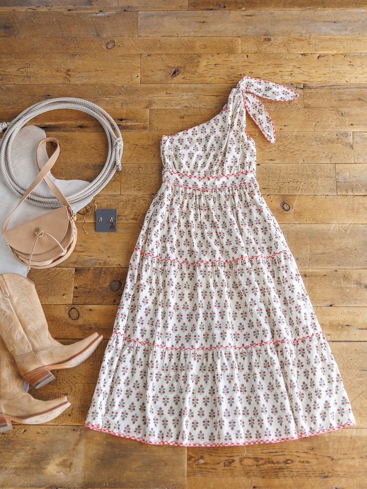 The Somerset Floral One Shoulder Dress by Savannah Sevens western life{&}style is styled on a wooden floor with tan cowboy boots, a beige handbag, a white rope belt, and earrings.