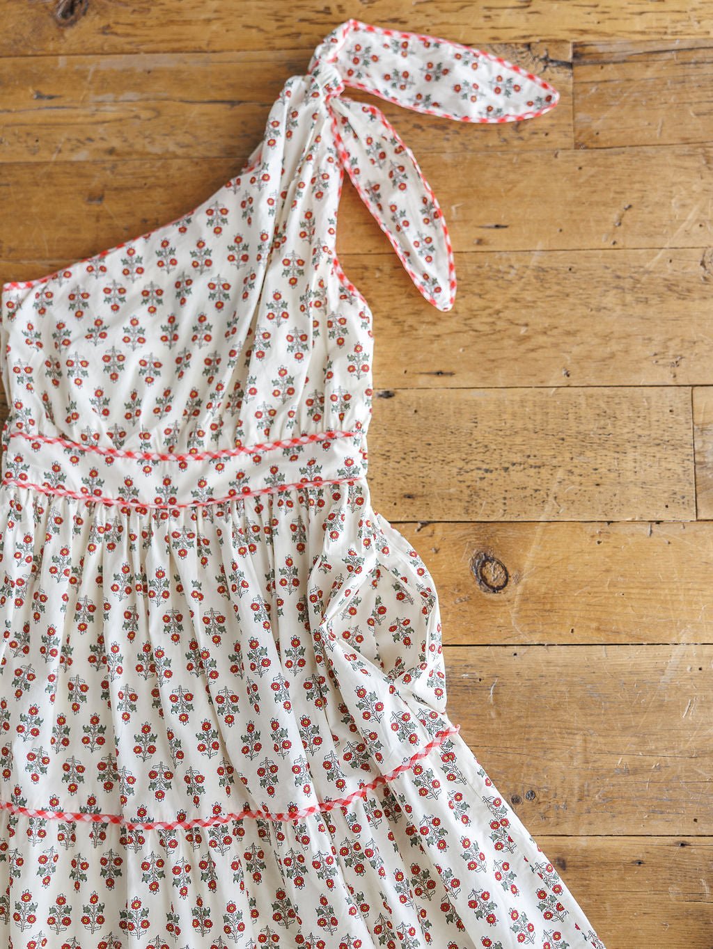 The Somerset Floral One Shoulder Dress by Savannah Sevens western life{&}style is a cream cotton midi dress with a cherry print, floral design, gingham trim, and a bow at the shoulder, shown laid flat on a wooden floor.