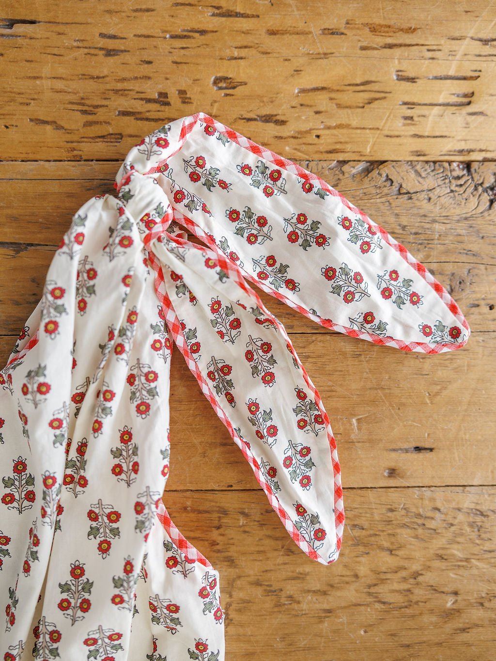 A detailed view of the Somerset Floral One Shoulder Dress by Savannah Sevens western life{&}style, featuring a red floral pattern and gingham trim, tied in a knot on rustic wood.