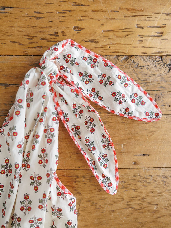 A detailed view of the Somerset Floral One Shoulder Dress by Savannah Sevens western life{&}style, featuring a red floral pattern and gingham trim, tied in a knot on rustic wood.