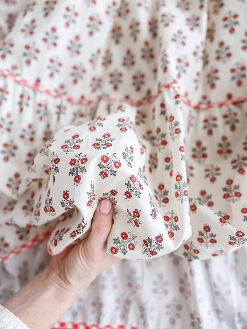 A hand displays the texture and detail of the Somerset Floral One Shoulder Dress by Savannah Sevens western life{&}style, featuring white cotton fabric with a red and green floral print and gingham trim against a matching background.