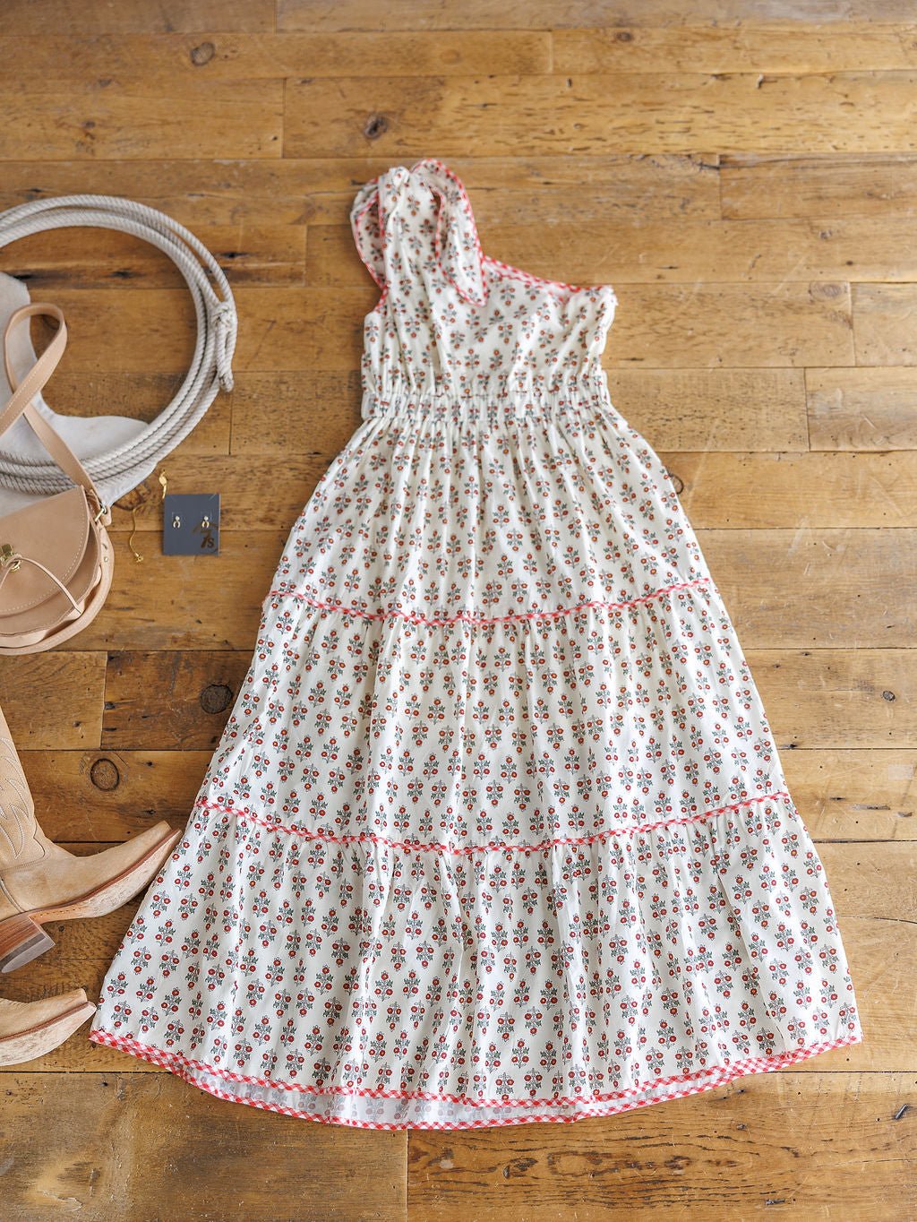 The Somerset Floral One Shoulder Dress by Savannah Sevens western life{&}style, featuring red floral patterns and a tiered skirt, is styled on a wooden floor with tan cowboy boots, a gray hat, beige purse, and earrings—perfect for warm days.