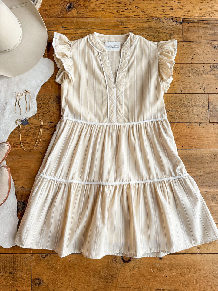 The Somerton Oatmeal Striped Dress by Savannah Sevens western life{&}style, featuring cream earth tones and ruffled shoulders, is displayed on wood with a white hat, gold jewelry, and a white cowhide rug.