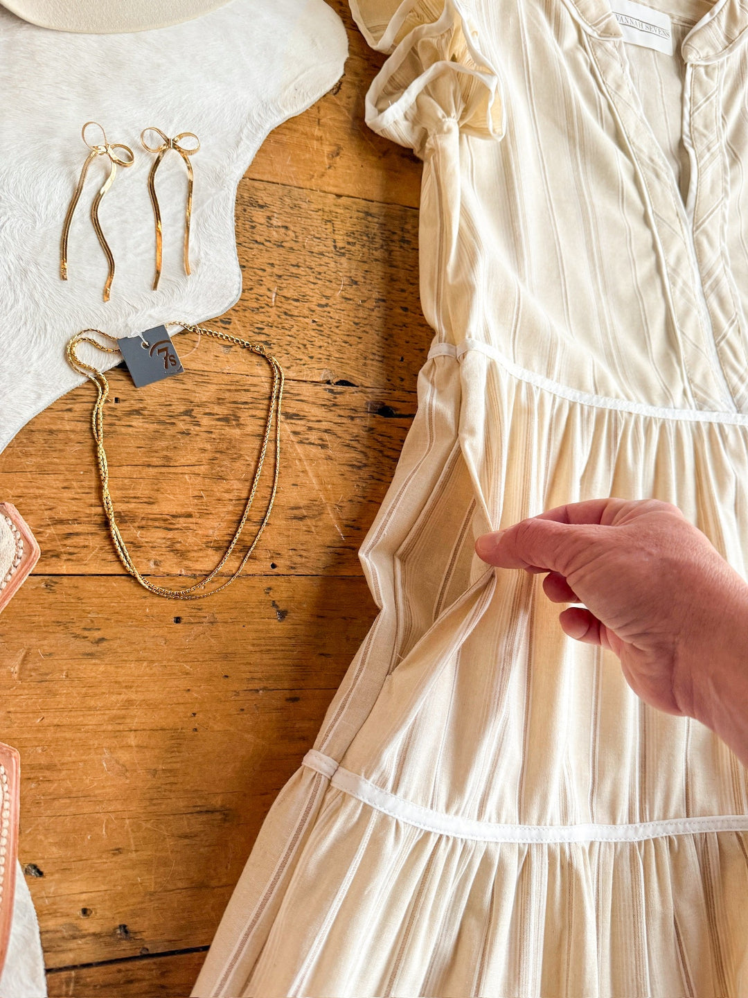 A hand touches the pocket of the Savanna Sevens western life{&}style Somerton Oatmeal Striped Dress, short-sleeved and laid on wood, with gold earrings, a gold necklace, earth-tone sandals, and part of a white hat arranged nearby.