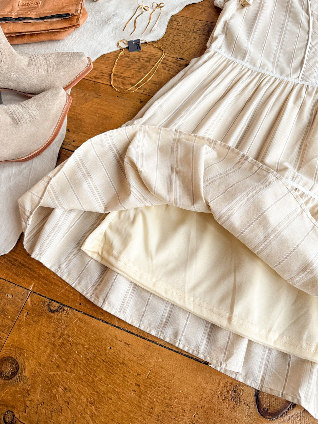 The Somerton Oatmeal Striped Dress by Savannah Sevens western life{&}style, featuring cap sleeves and a ruffled hem, is displayed on wood with beige boots, a brown purse, gold jewelry, and a white cloth. The dress is slightly lifted to show its lining.