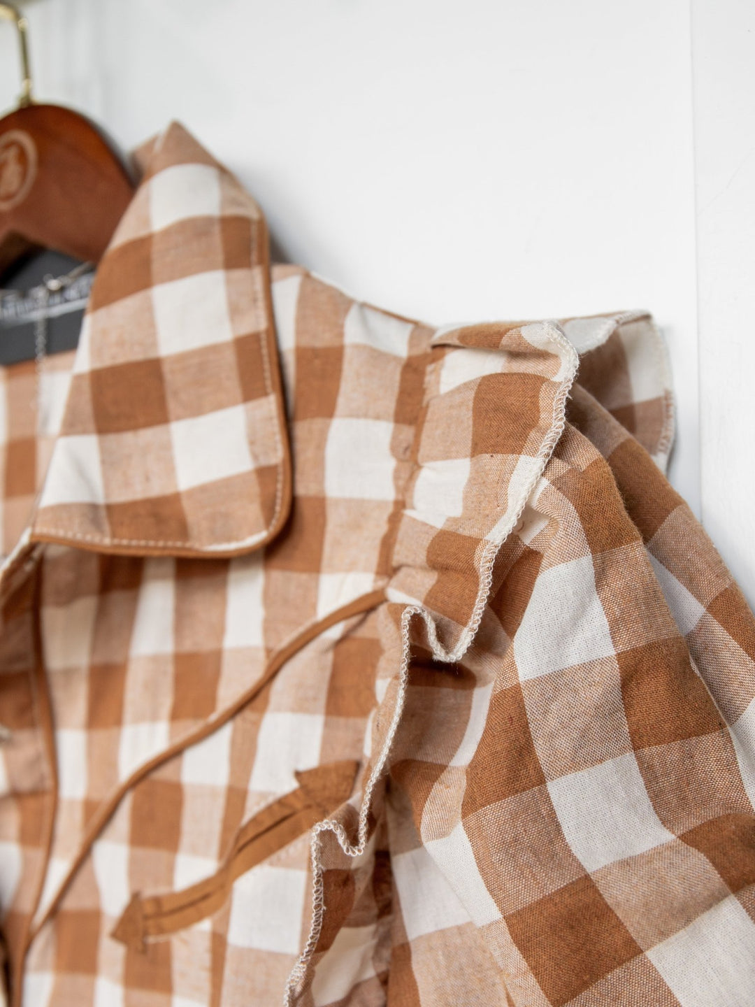 Close-up of the Somona Brown Gingham Pearl Snap Top by Savannah Sevens western life{&}style, featuring ruffled trim on the shoulders and a pointed collar, hanging on a hanger against a white background.
