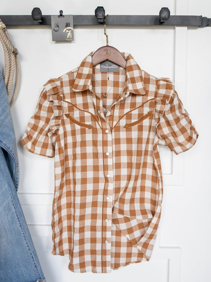 The Somona Brown Gingham Pearl Snap Top by Savannah Sevens western life{&}style, featuring short sleeves, hangs on a wooden hanger with blue jeans partially visible to the left.