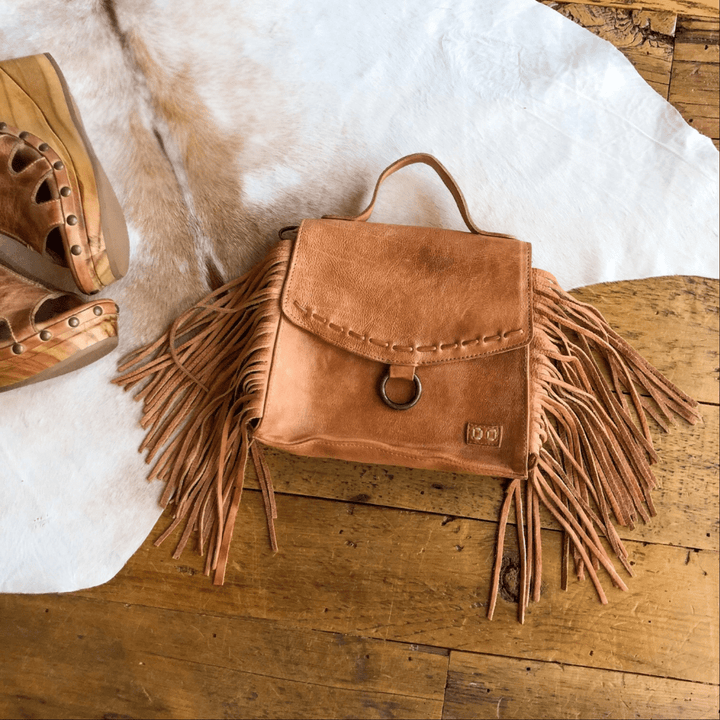 The Sonora Fringe Leather Crossbody by Savannah Sevens western life{&}style, a tan leather bag with fringe sides and a metal ring flap, rests on a white cowhide rug beside wooden clogs on a wooden floor. By BedStu. 