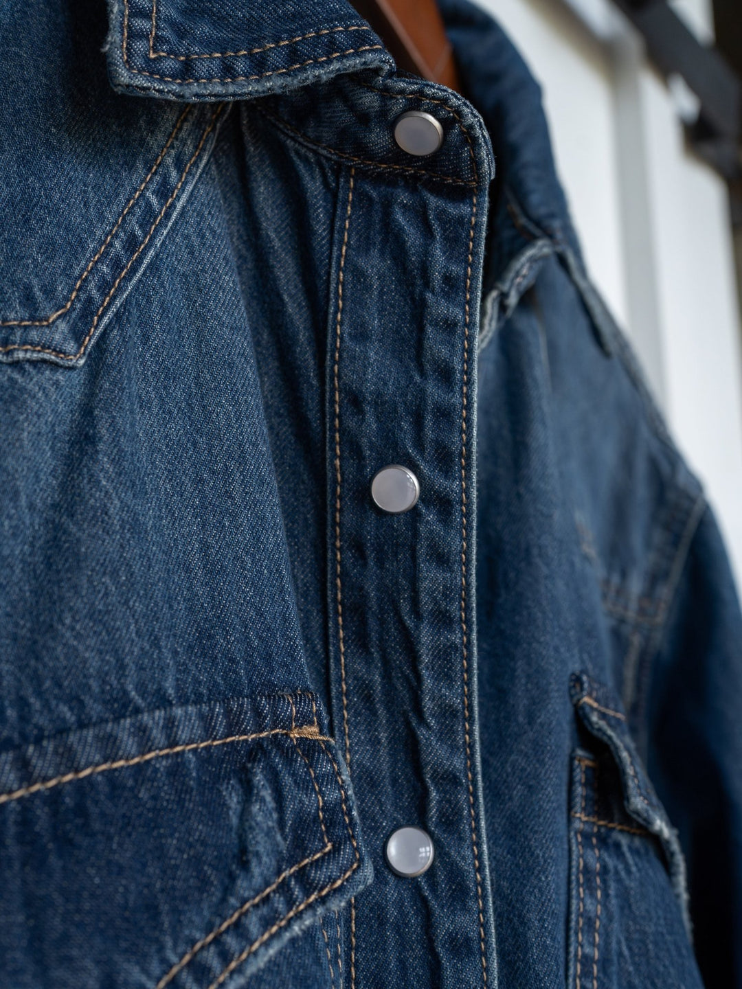 Close-up of The Sorento denim shirt by Savannah Sevens western life{&}style, featuring blue distressed fabric, western yoke, visible stitching, detailed pockets, and silver snap buttons along the front. Texture and craftsmanship are highlighted.