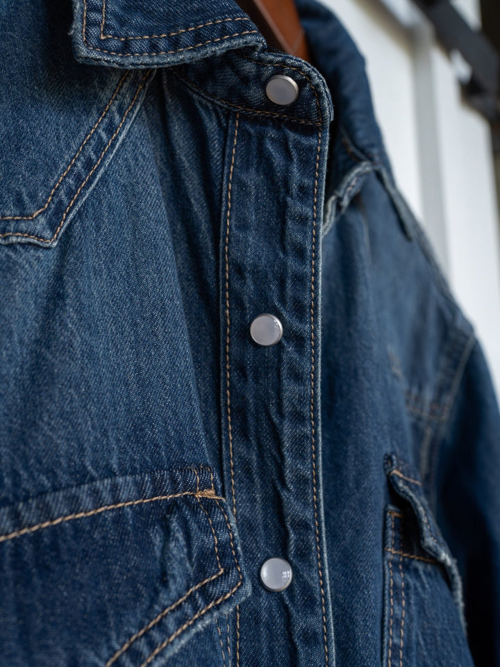 Close-up of The Sorento denim shirt by Savannah Sevens western life{&}style, featuring blue distressed fabric, western yoke, visible stitching, detailed pockets, and silver snap buttons along the front. Texture and craftsmanship are highlighted.